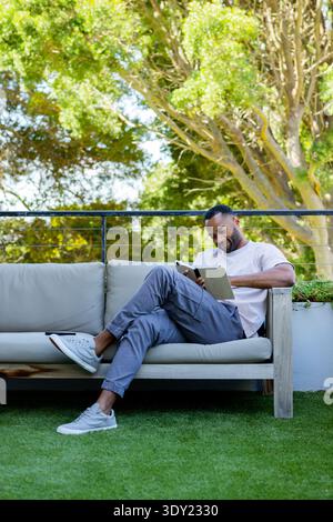African American mature man sitting on patio sofa holding tablet smartphone on cushion, copy space. Adult, lifestyle, minimalist, terrace, outdoor, gr Stock Photo