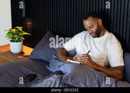 Middle-aged African American man writing in notebook on bed at home with pen, smartphone, pillows. Journal, serene, cozy, bedroom, minimalist, ambient Stock Photo