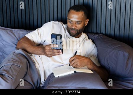 Mature African man reclining on bed using smartphone writing in notebook with pen in tan tee. Bedroom, headboard, bedding, phone, journal, notepad, ca Stock Photo