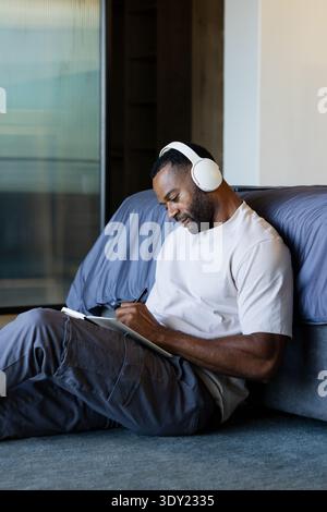 African American man sitting on bedroom floor leaning by bed writing in notebook with white headset. Mature, focused, headphones, journaling, solitary Stock Photo