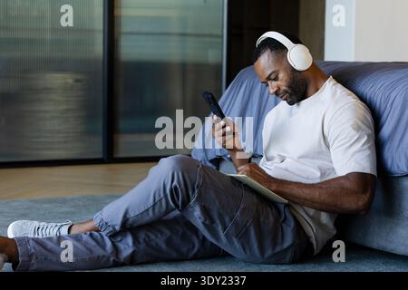 African American man sitting on rug by low bed wearing headphones, checking phone, writing notebook. Tablet, focus, modern, home, calm, lifestyle, stu Stock Photo