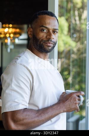 African American man standing at home by glass door holding white mug, chandelier glowing behind. Portrait, lifestyle, warmth, softlight, window, gree Stock Photo