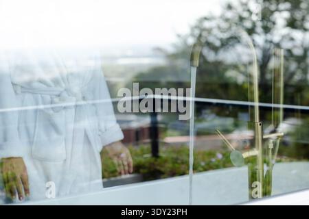 Adult African American woman standing at glass in bath wearing robe, brass tap running, copy space. Reflections, terrace, balcony, greenery, daylight, Stock Photo