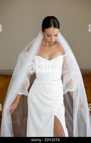 Bride wearing an elegant white wedding dress and veil, gazing down thoughtfully, embodying beauty and romance in a serene indoor setting Stock Photo