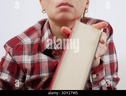 Thoughtful Female Student in Plaid Shirt Pressing Textbook Against Cheek Looking Forward in Deep Thought During University Study and Learning Stock Photo