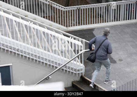 Senior man ascending urban stairs at right, holding metal handrail and crossbody bag, copy space. Cityscape, staircase, railing, pavement, wet, concre Stock Photo