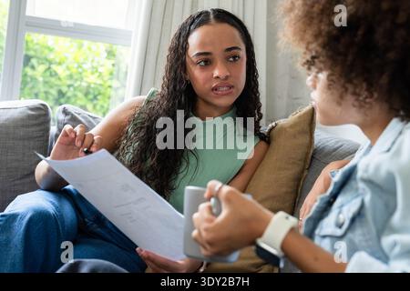 Female friends sitting on sofa discussing document, other holding mug, wearing mint top smartwatch. Women, discussion, home, cozy, natural, daylight, Stock Photo