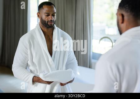 Mature African American man standing in bathroom wearing bathrobe holding white towel facing mirror. Serene, private, daylight, minimalist, tub, chrom Stock Photo