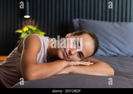 Female lying on bed in bedroom resting head on arms with lit lamp and plant nearby. Repose, serene, intimate, softlighting, slats, bedside, contempora Stock Photo