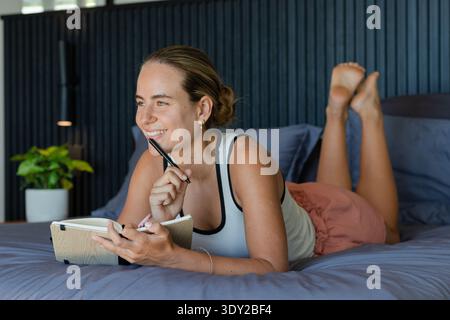 Adult woman in white tank top lying on bed, holding pen, open notebook, smiling, looking left. Bedroom, female, journal, writing, minimal, natural, li Stock Photo