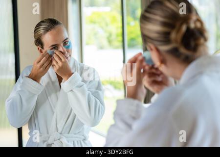 Female standing at mirror in home bathroom applying blue under-eye gel patches wearing white robe. Skincare, selfcare, eyemask, daylight, glow, minima Stock Photo