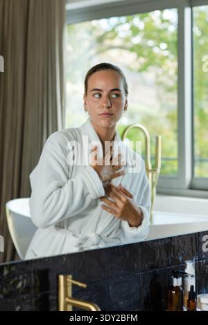 Woman standing at bathroom vanity adjusting white bathrobe beside window with freestanding bathtub. Robe, tub, marble, brass, daylight, glass, greener Stock Photo