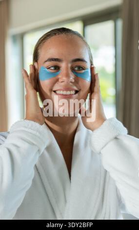 Female smiling and adjusting blue hydrogel eye patches at home near large window in bathrobe. Selfcare, skincare, pampering, routine, morning, glow, s Stock Photo