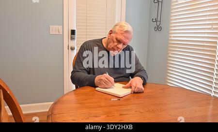 Senior man writes notes at wooden dining table. Calm focus reflects quiet concentration. Soft natural light enhances peaceful atmosphere. Ideal for th Stock Photo