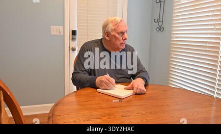 Man sits at wooden table, writing in notebook. He concentrates, eyes focused on paper. Soft light from window filters through blinds. Quiet indoor set Stock Photo