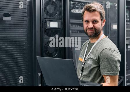 Smiling IT technician working on laptop in a server room surrounded by racks and equipment Stock Photo