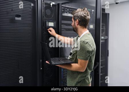 IT technician configuring server equipment in a data center while holding a laptop and checking network settings Stock Photo