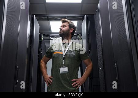 Data center technician inspecting server racks in a modern server room with confident posture and ID badge Stock Photo
