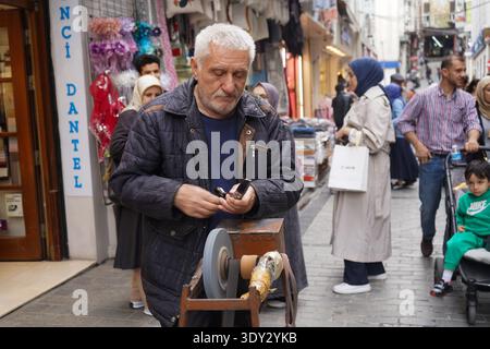 An elderly man meticulously sharpens a knife using a traditional manual grinding wheel on a city sidewalk. Stock Photo