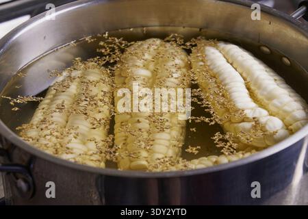 White corn cobs cooking in a pot on the stovetop, seasoned with aromatic spices Stock Photo