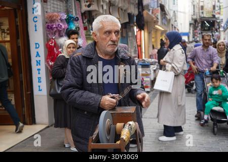 An elderly man meticulously sharpens a knife using a traditional manual grinding wheel on a city sidewalk. Stock Photo