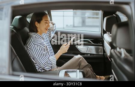 A Korean woman sits in the back seat of a car. She looks at her phone and smiles while the sunlight shines through the window. The city can be seen in Stock Photo