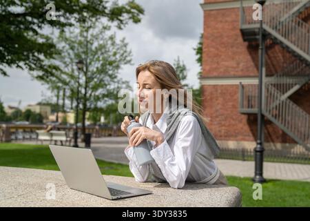 Relaxed businesswoman enjoying remote work outdoors, taking part in online meeting on computer. Stock Photo