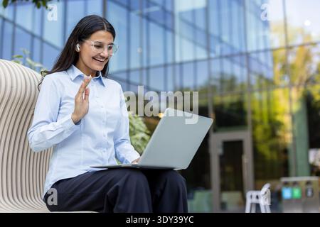 Young woman smiling and waving during a video call on her laptop, wearing earbuds and glasses, working remotely from an outdoor bench in front of a modern office building Stock Photo