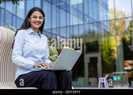 Young professional woman smiling, using a laptop and wearing earbuds while sitting on a modern bench outside a corporate office building, symbolizing remote work and technological connectivity Stock Photo