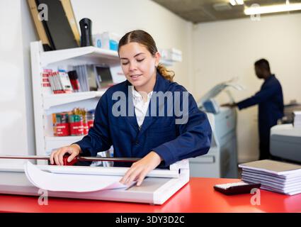 Portrait of cheerful young female typographer in uniform using paper cutter during work in printer shop Stock Photo