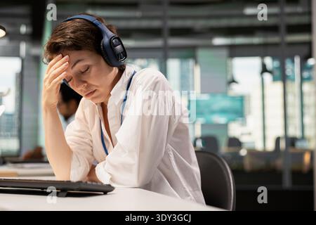 Tired programmer in Silicon Valley company office listening music to destress. Exhausted IT specialist wearing headphones closing eyes to relax at work while debugging applications Stock Photo