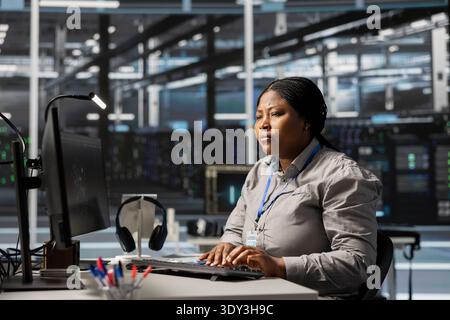 Data center software developer uses PC to update servers tech, configuring gear using program. Woman working in server farm reviewing hardware analytics, ensuring reliable network operations Stock Photo