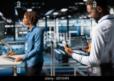 Server room IT programmer visualizing large language model infrastructure using tablet on elevated industrial platform. Data center system administrators collaborating on tasks. Stock Photo