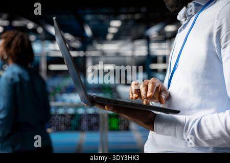 Close up of data center software engineer doing maintenance work on laptop, ensuring optimal performance. Male worker reviewing diagnostics and debugging server hub high tech equipment. Stock Photo