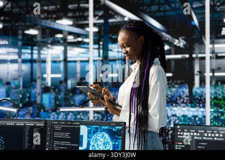Technician using tablet in data center, configuring hardware specifically optimized for AI tasks. African american woman in server farm automating system diagnostics using machine learning predictions Stock Photo