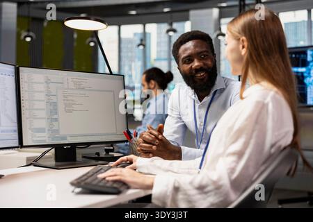Happy senior programmer mentoring junior developer at desk in office, teaching best coding practices. Cheerful tech company lead engineer guiding intern through project tasks Stock Photo
