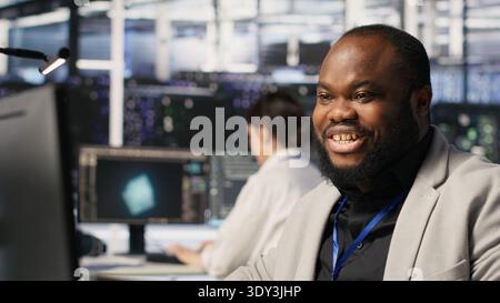 Data center system administrator monitoring servers gear performing AI training and inference tasks. Man working in server room oversees computing systems, managing deep learning operations, camera A Stock Photo