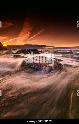 A breathtaking long exposure seascape capturing the dynamic movement of ocean waves washing over dark coastal rocks during a vibrant sunset. The sky i Stock Photo
