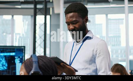 Technician next to office coworkers developing software applications, integrating artificial intelligence automation. African american man next to developers at work debugging applications, camera A Stock Photo