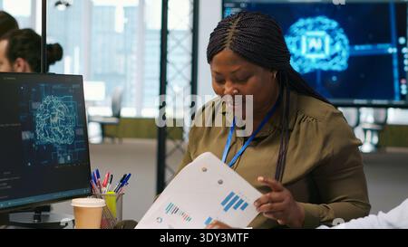 Joyous technician with disability and coworker chatting while coding AI algorithms debugging software issues. Upbeat tech startup office IT expert in wheelchair and colleague using computer, camera A Stock Photo