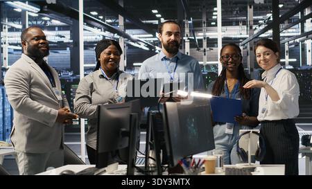 Smiling engineering team in server room reviewing infrastructure diagnostics on devices. IT professionals in data center analyzing performance data to maintain system integrity and uptime, camera A Stock Photo