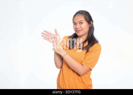 Portrait of a smiling Indonesian woman in orange shirt using sign language to perform the gesture for "Name", isolated on a plain white background. Stock Photo