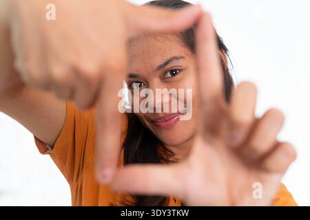 Portrait of a happy Indonesian woman in orange shirt using fingers to create a frame around her face, isolated on a plain white background. Stock Photo