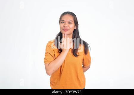 Young Indonesian woman in orange shirt using sign language to say thank you, smiling and gesturing with her hand near her chin. Stock Photo
