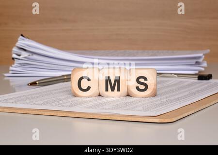 Front view of three wooden blocks spelling the acronym CMS placed on a clipboard with a financial document and a pen. Stock Photo