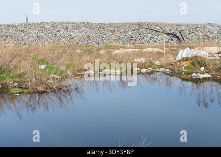 Trash is piled up in the background while water sits in the foreground surrounded by dry grass and small plants under clear blue skies. Stock Photo