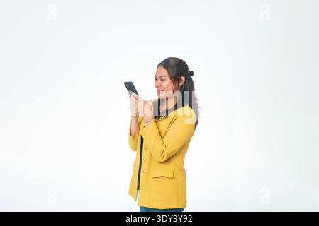 Side profile of Indonesian woman in yellow blazer looking down at smartphone screen with focused expression, isolated on white background. Stock Photo