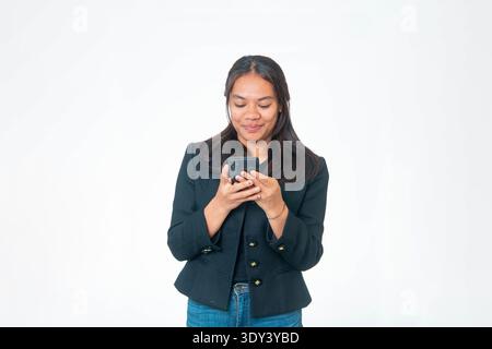 Indonesian woman wearing black textured jacket and blue jeans, holding black smartphone with both hands while looking down at screen Stock Photo