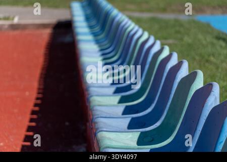 Plastic seats are arranged in rows along a sports field. The sun shines down, highlighting the empty seats and green grass nearby. Stock Photo