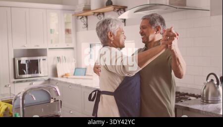 Dancing senior couple holding hands in home kitchen, wearing apron and olive-green shirt, kettle Stock Photo